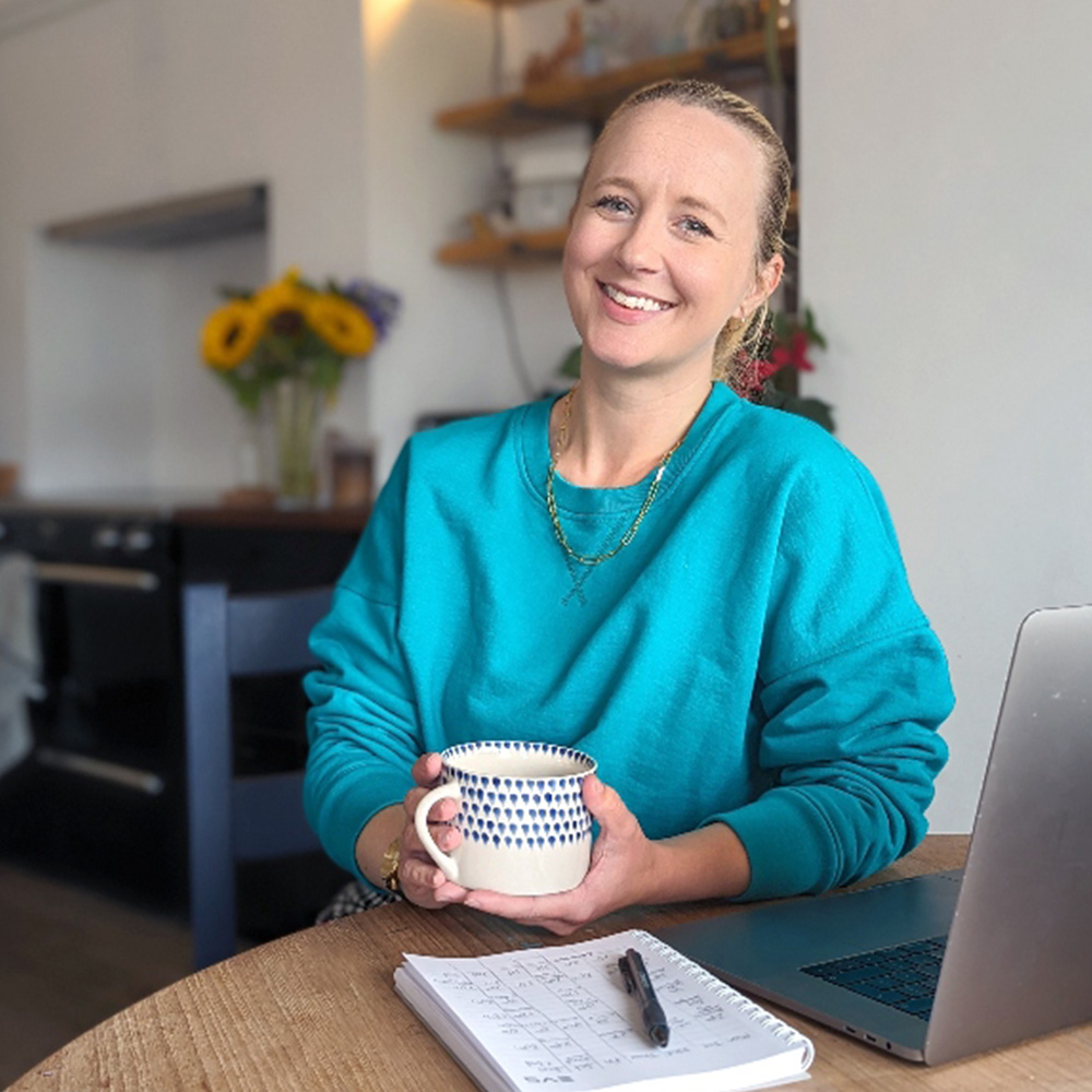 Lindsay Wright the Freelance Writer and Designer from Lancashire wearing a blue jumper holding a cup of tea whilst sat at a laptop.